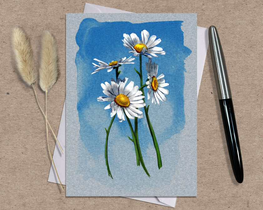 Greeting card with daisy flowers on a blue watercolor background, placed on a textured surface with a pen and dried plants.