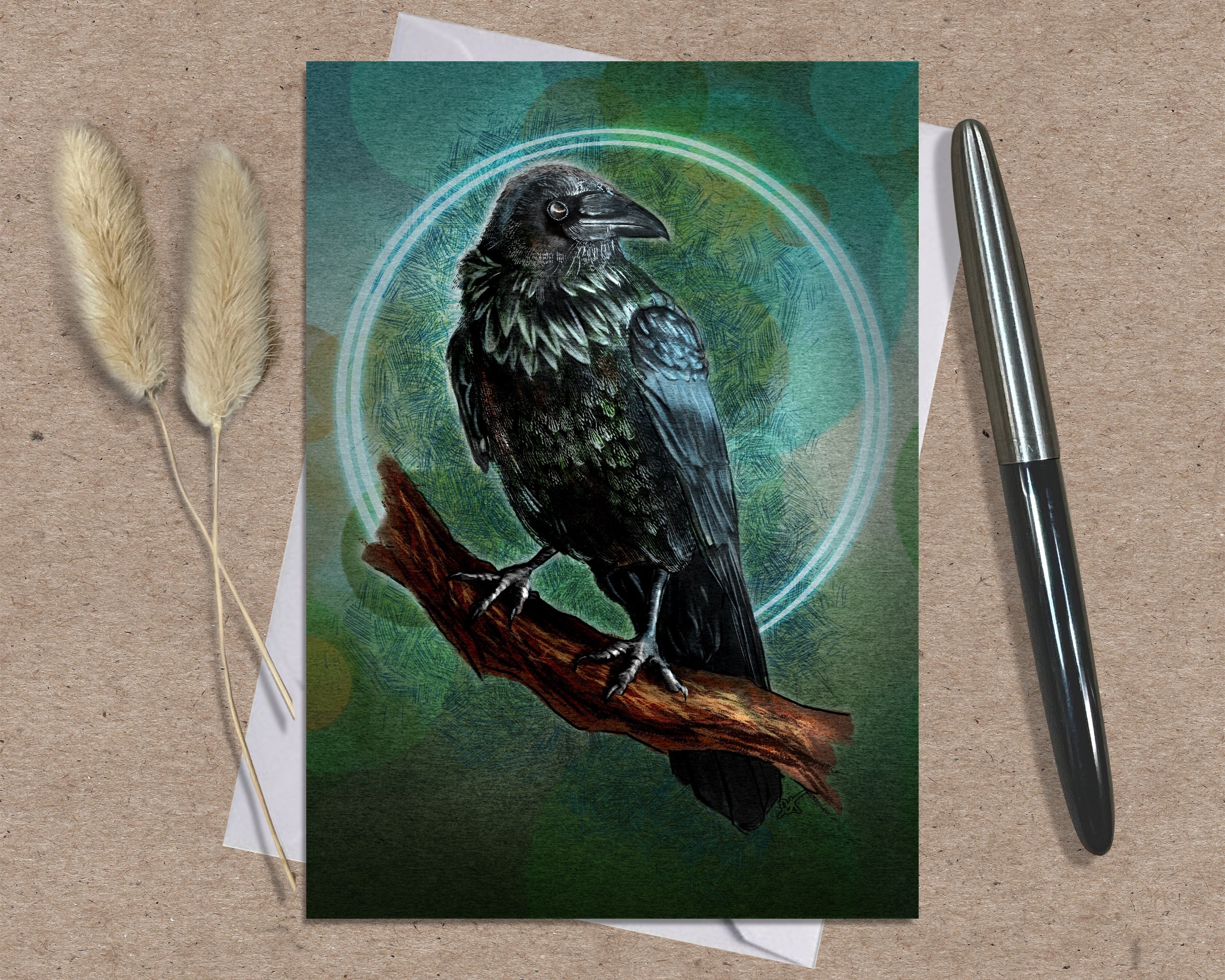 Greeting card with a crow illustration on a branch, placed on a textured surface with a pen and dried plants.