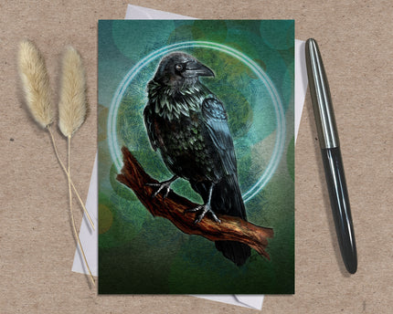 Greeting card with a crow illustration on a branch, placed on a textured surface with a pen and dried plants.