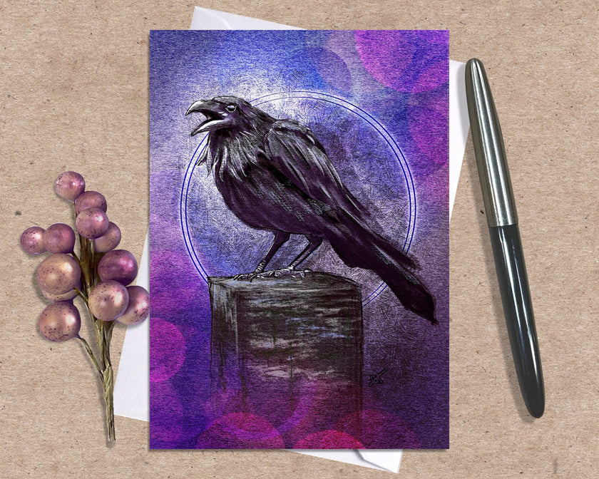 Greeting card with a raven illustration on a purple and blue gradient background, placed on a textured surface with a pen and decorative elements.