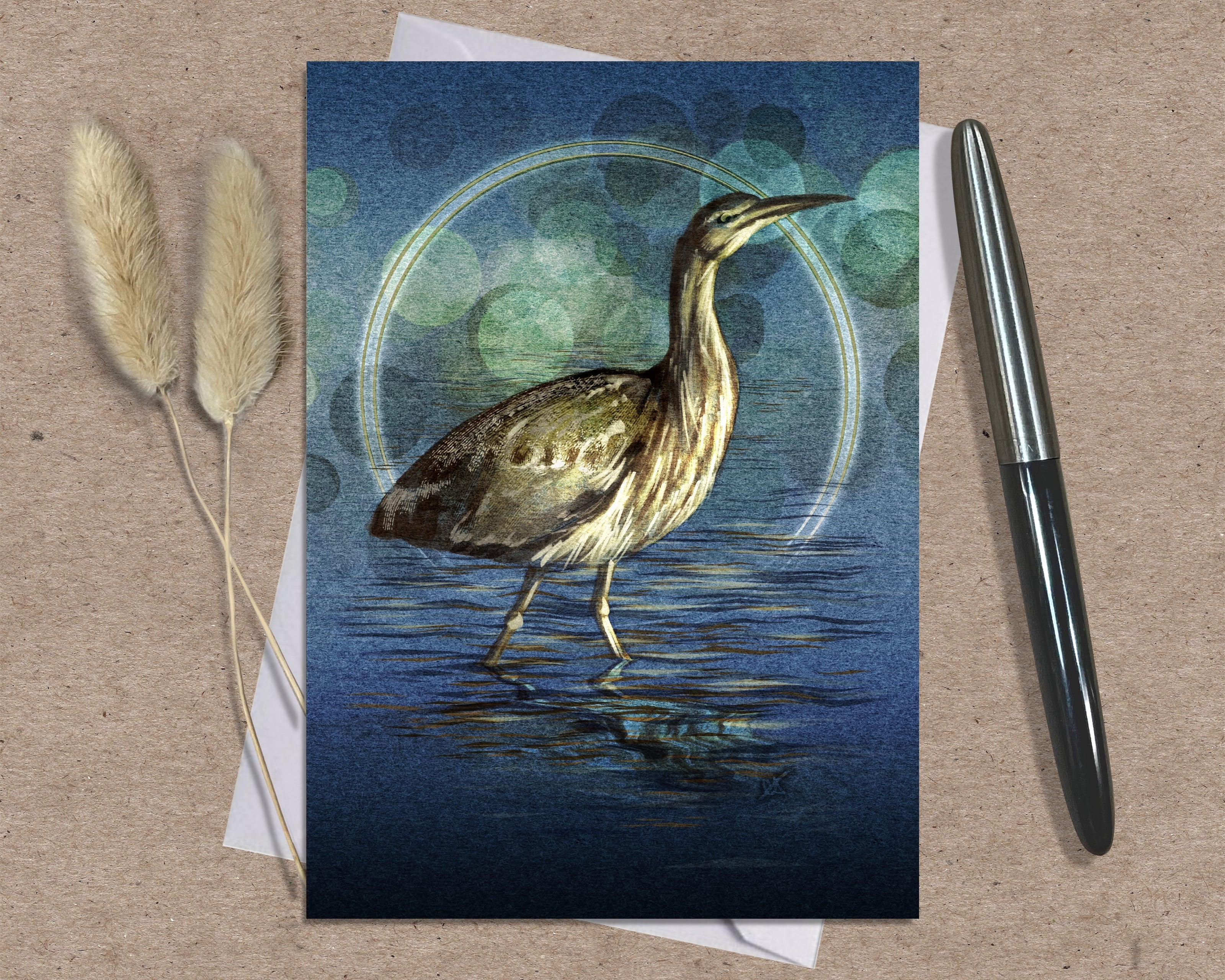 Greeting card with a bittern design on a textured surface with a pen and dried plants.