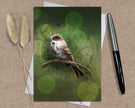 Card with a bird illustration on a green background, placed on a textured surface with a pen and dried plants.