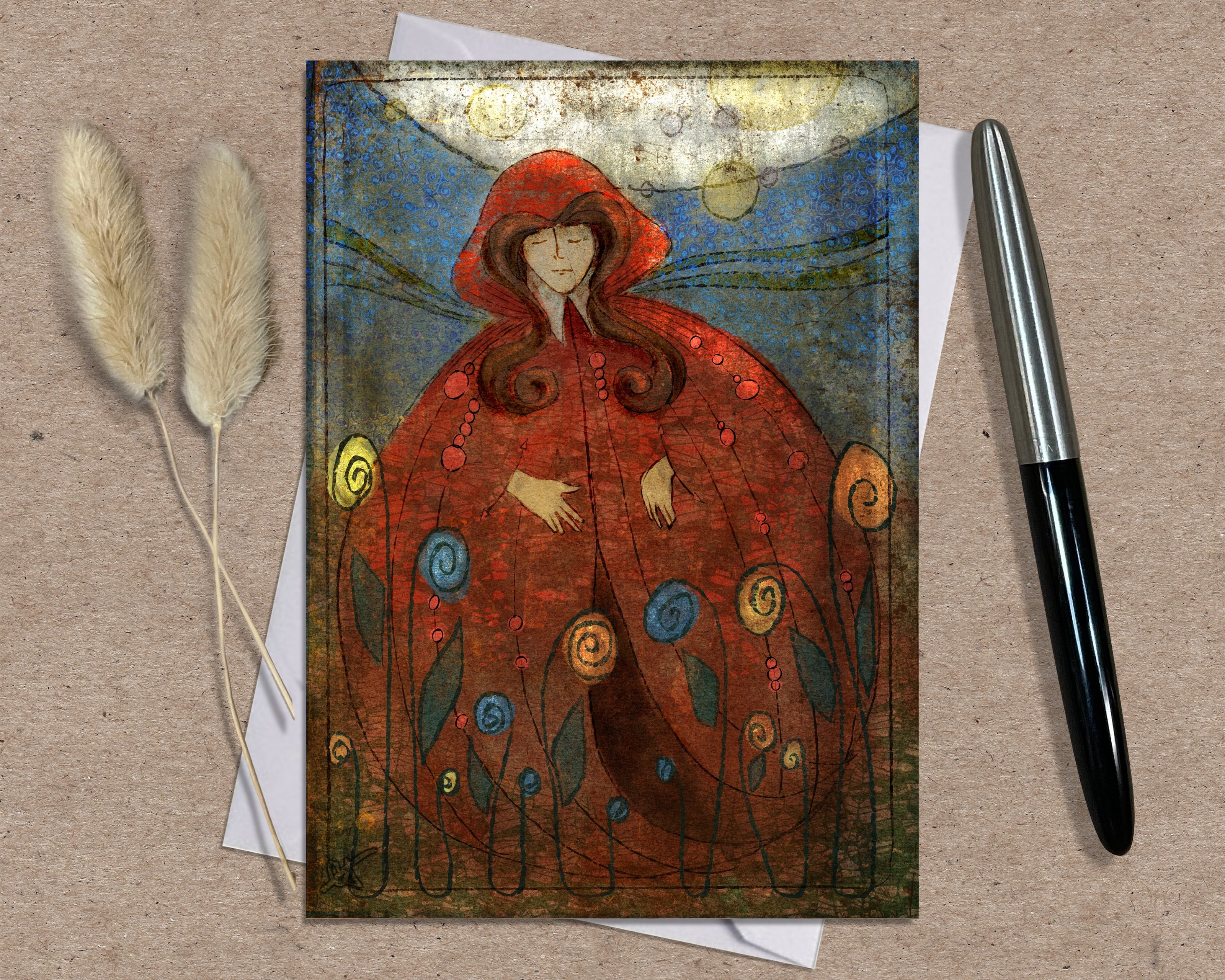 Greeting card with a red hooded figure on a textured surface with a pen and dried plants.