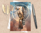 Greeting card with a hawk illustration on a textured surface with a pen and dried plants.
