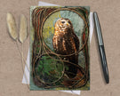 Greeting card with an owl design on a textured surface with a pen and dried plants.