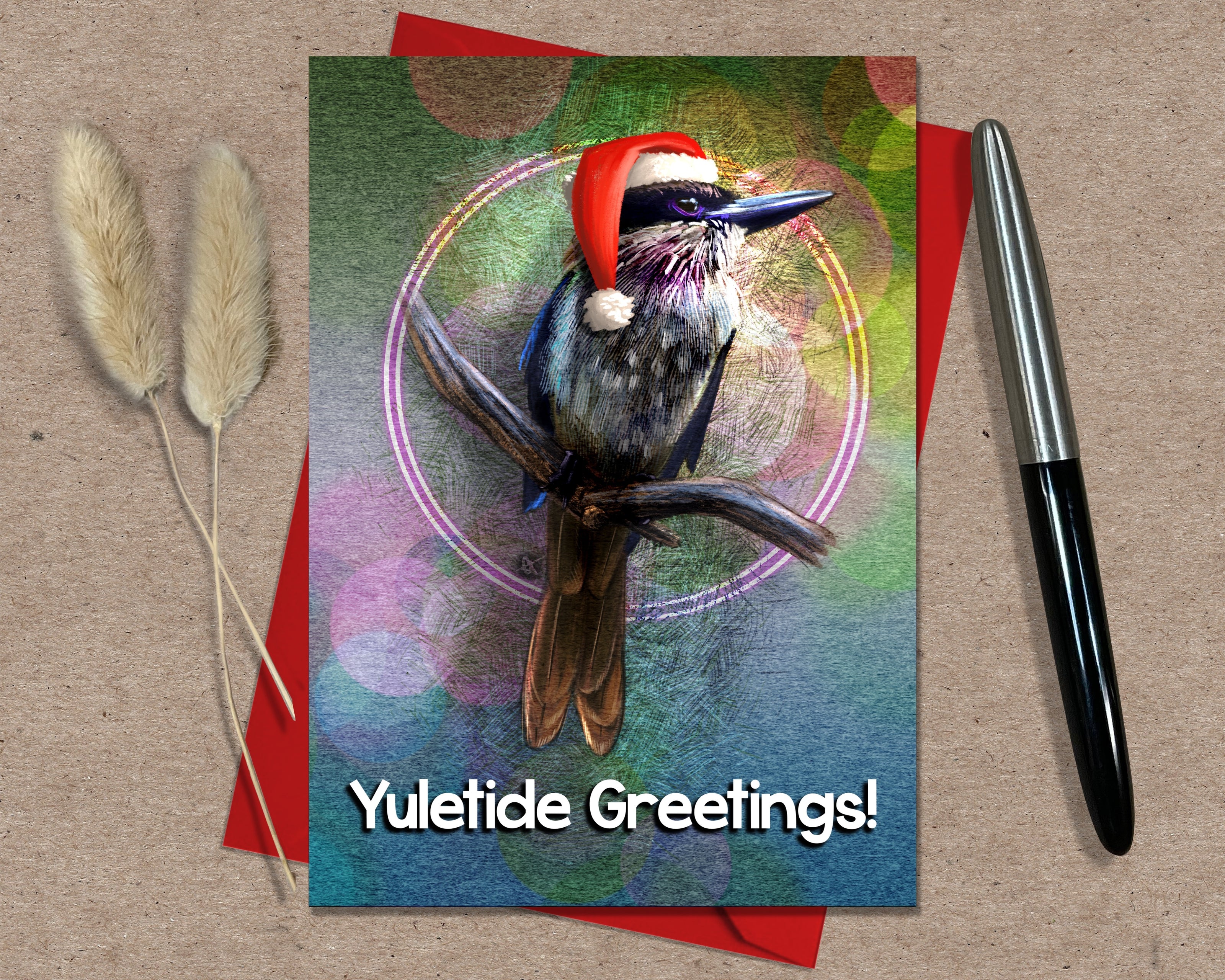 Greeting card with a bird wearing a Santa hat and 'Yuletide Greetings!' text, placed on a textured surface with a pen and dried plants.