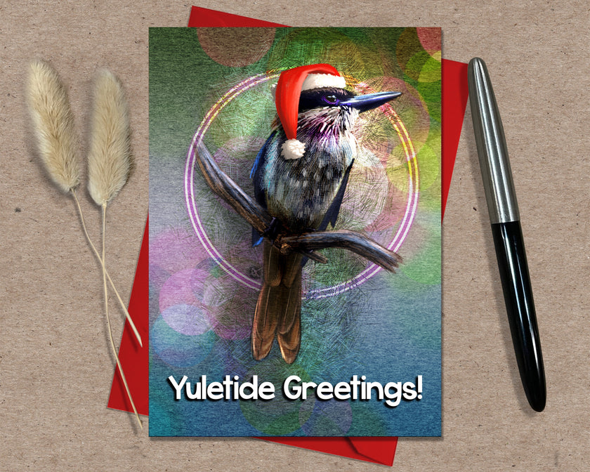 Greeting card with a bird wearing a Santa hat and 'Yuletide Greetings!' text, placed on a textured surface with a pen and dried plants.