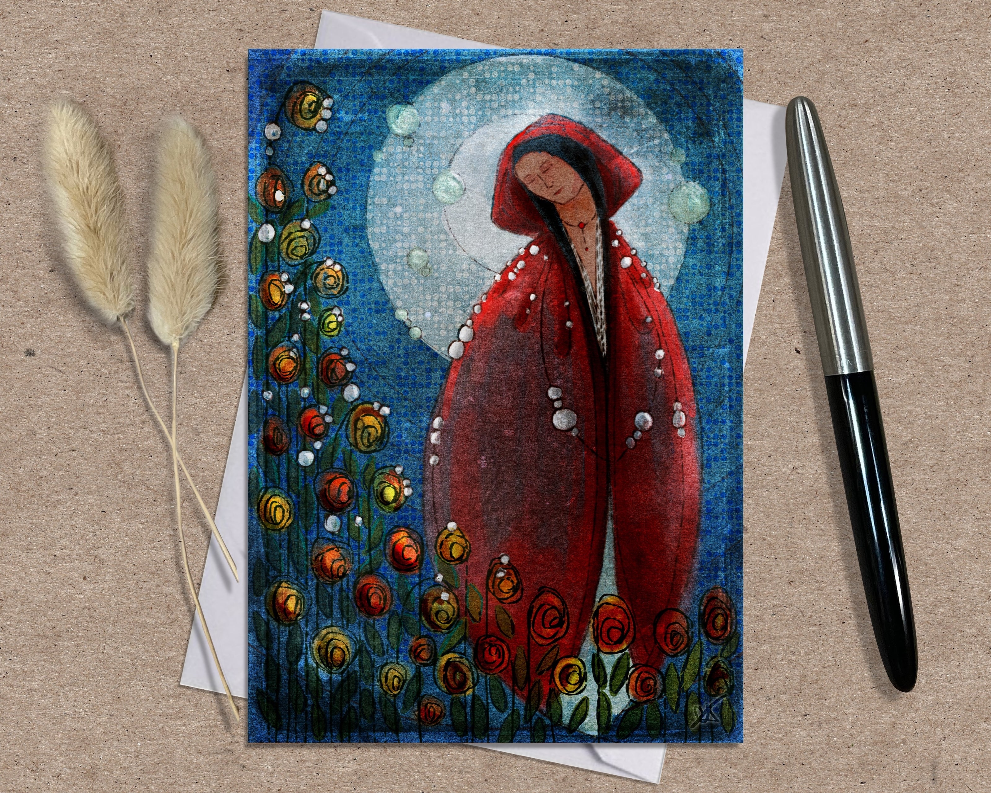 Greeting card with a red-robed figure surrounded by flowers on a textured surface with a pen and dried plants.