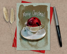 Christmas card with a red bell in a teacup, 'Merry Christmas!' text, on a textured surface with a pen and dried plants.
