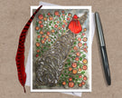 Greeting card with a stylized tree design featuring flowers and a person in a red cloak placed on a textured surface with a pen and feather.