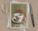 Greeting card with a teacup design on a textured surface with a pen and dried plants.