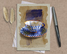 Greeting card with a blue and white teacup design on a textured surface with a pen and dried plants.