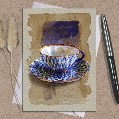 Greeting card with a blue and white teacup design on a textured surface with a pen and dried plants.