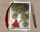 Greeting card with a design of a red-robed figure and leaves, accompanied by a pen and feather on a textured surface.