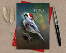 Christmas card with a bird wearing a Santa hat and 'Peace and Joy' text, placed on a textured surface with a pen and dried plants.