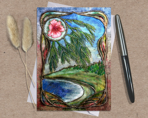 Decorative card with nature-themed design featuring a flower, trees, and water on a textured surface with a pen and dried plants.