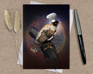 Card with a bird wearing a chef's hat on a wooden branch, placed on a textured surface with a pen and dried plants.
