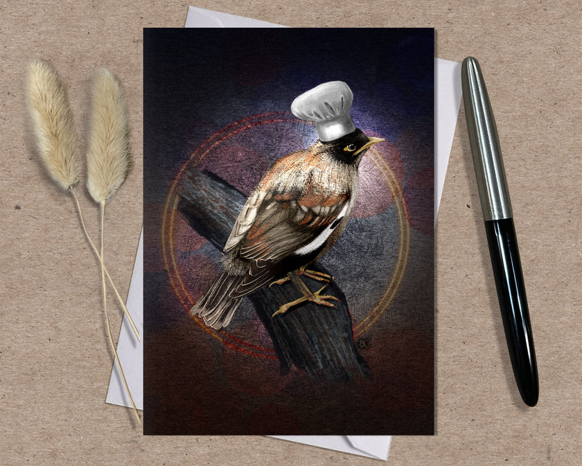 Card with a bird wearing a chef's hat on a wooden branch, placed on a textured surface with a pen and dried plants.