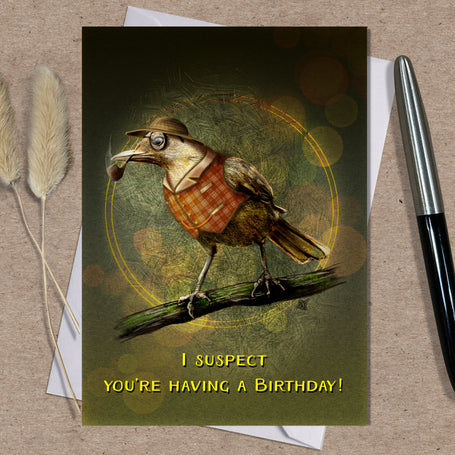 Birthday card with a bird illustration and text on a textured surface with a pen and dried plants. Text on the card says "I suspect you're having a birthday!"