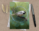 Greeting card with a bird illustration on a green background, accompanied by dried plants and a pen.